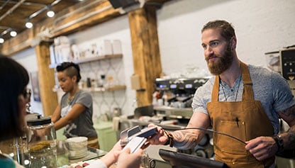 Woman paying for a beverage in a coffee shop, exchanging payment with a smiling bearded man