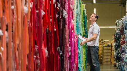 Man inspecting rolls of colorful fabric in a warehouse, holding a clipboard