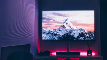 A modern flat-screen TV displaying a mountain scene, illuminated by pink and purple LED lighting.