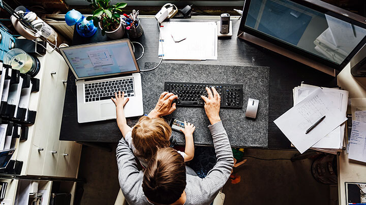 Man and child sitting at desk while man types on a laptop and child plays on a phone