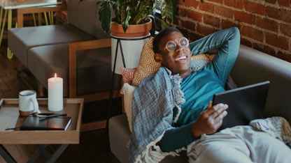 A man relaxes on a couch with a tablet, wrapped in a blanket, next to a lit candle and mug.