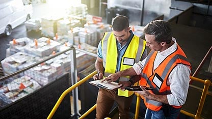 Two men in safety vests discussing data points in a notebook