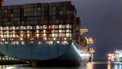 A large cargo ship loaded with containers is docked at night, illuminated by port lights.