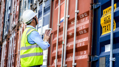 Inspector in safety gear examines shipping containers using a tablet at a port