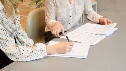 Two people reviewing and discussing documents at a table, one pointing and the other taking notes.